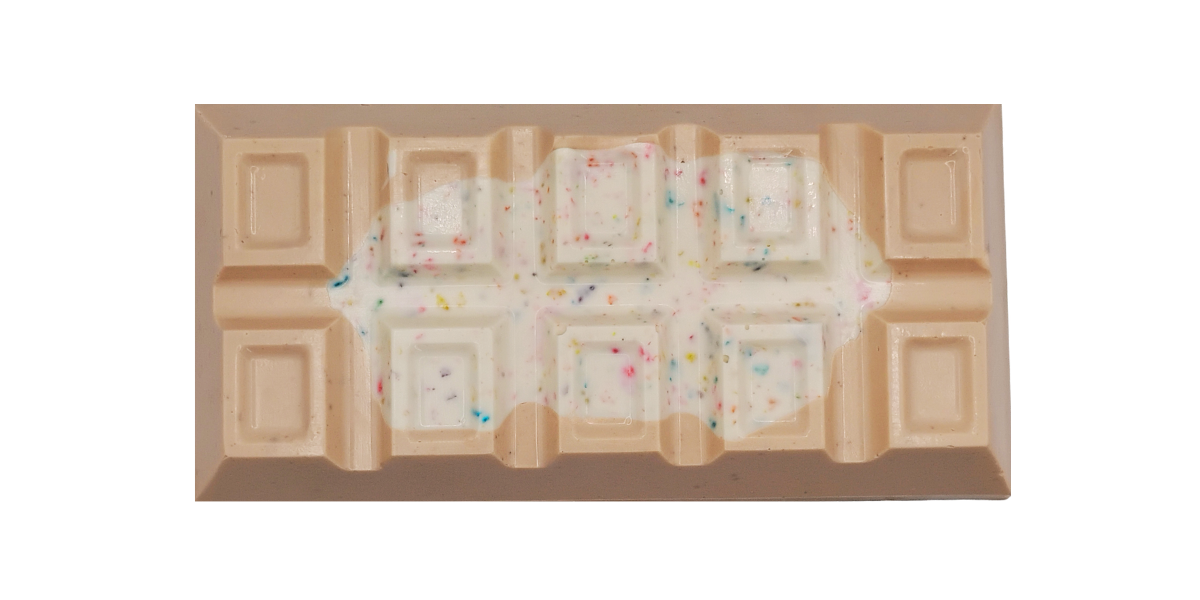 Rectangular tan chocolate bar with raised square segments, topped with a white chocolate layer and scattered multicolored birthday color specs across the center
